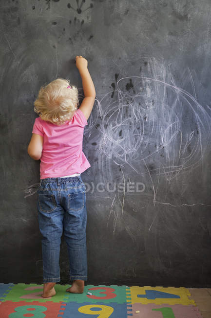 Rear view of cute blonde little girl writing on a blackboard — Stock Photo