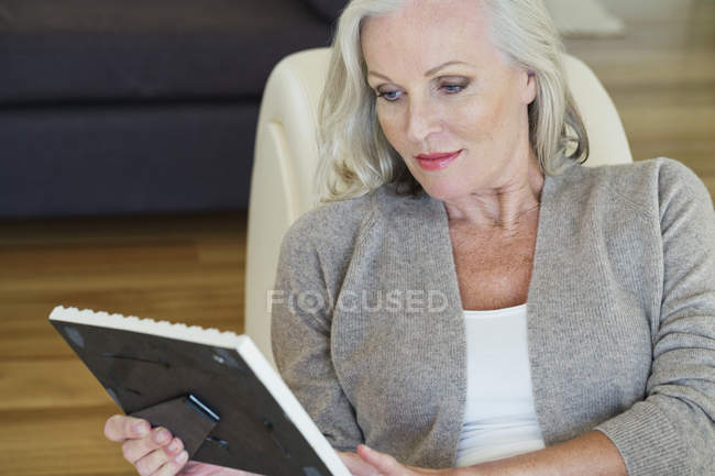 Пожилая женщина смотрит на рамку картины, сидя дома на диване — стоковое фото