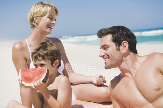 Niño comiendo una sandía con los padres en la playa - foto de stock