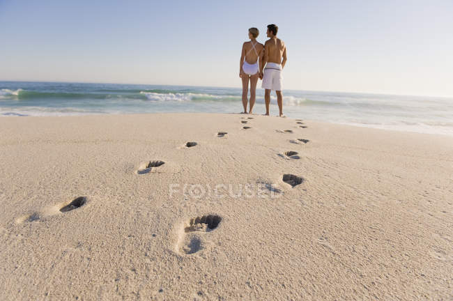 Huellas en la playa de arena con la pareja de pie sobre el fondo y mirando a la vista - foto de stock