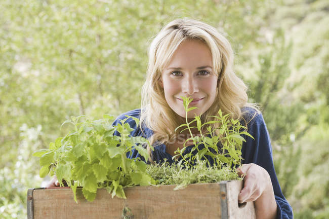 Porträt einer jungen blonden Frau, die eine Kiste mit Pflanzen im Garten trägt — Stockfoto