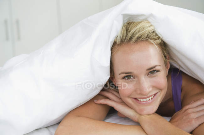 Nahaufnahme einer Frau, die auf dem Bett unter einer Bettdecke liegt und lächelt — Stockfoto