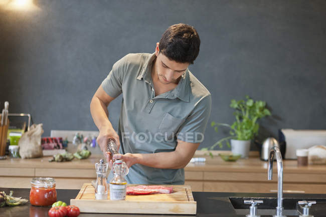 Man sprinkling black pepper on meat in modern kitchen — Stock Photo