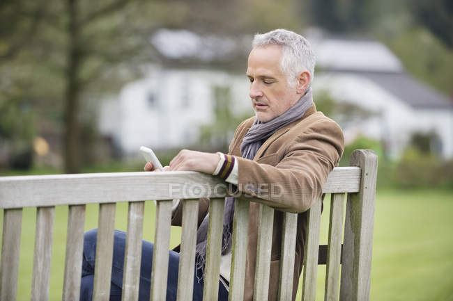 Homem maduro elegante usando telefone celular no banco de madeira no parque — Fotografia de Stock