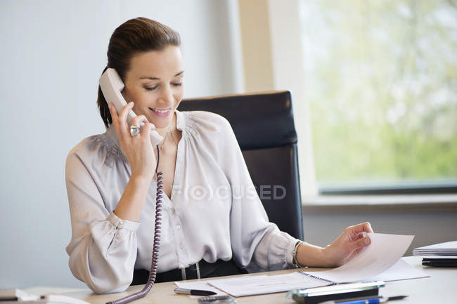 Усміхнена бізнес-леді розмовляє по стаціонарному телефону в офісі — стокове фото