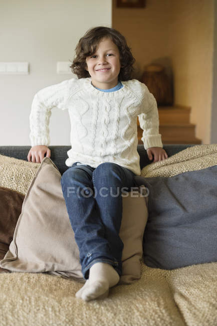 Retrato de niño sonriente sentado en almohadas en el sofá en casa - foto de stock