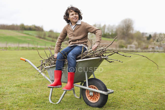 Garçon assis sur brouette avec du bois de chauffage dans un champ — Photo de stock