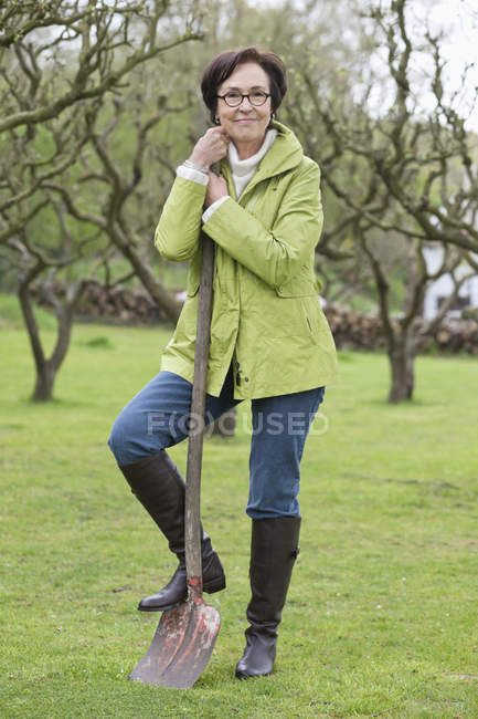 Пожилая женщина, стоящая с лопатой в саду и смотрящая в камеру — стоковое фото