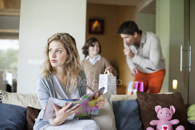 Woman sitting on couch with her husband and son holding surprise gifts in background at mother day — Stock Photo