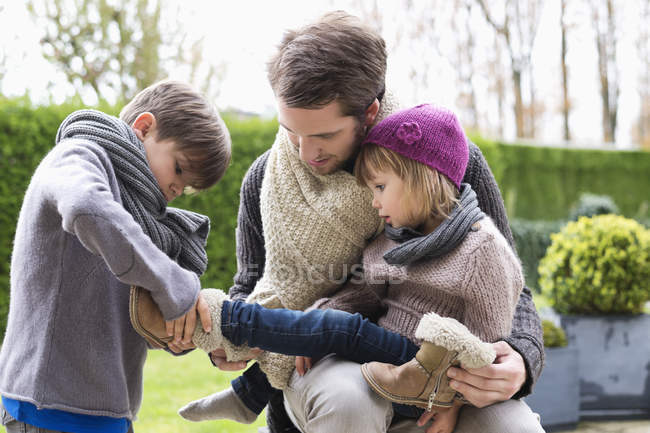 Chico poniendo en zapatos en hermana sentado en padre regazo al aire libre - foto de stock