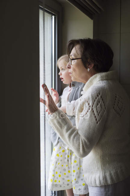 Mulher com pequena neta olhando através da janela — Fotografia de Stock
