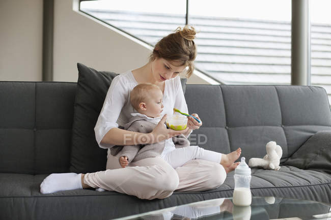Frau füttert kleine Tochter, während sie zu Hause auf dem Sofa sitzt — Stockfoto