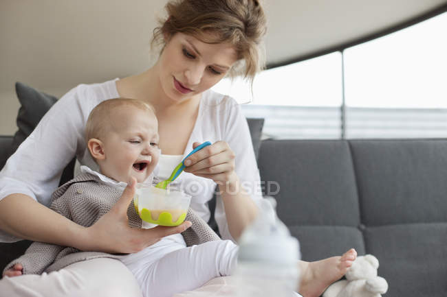 Junge Frau füttert kleine Tochter zu Hause auf Sofa — Stockfoto