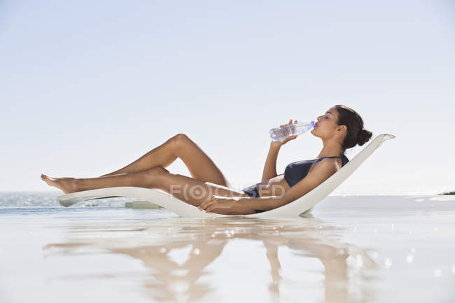Donna magra rilassata in costume da bagno acqua potabile sulla sedia a sdraio sulla spiaggia — Foto stock