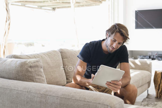 Усміхнений молодий чоловік сидить на дивані і використовує цифровий планшет — стокове фото