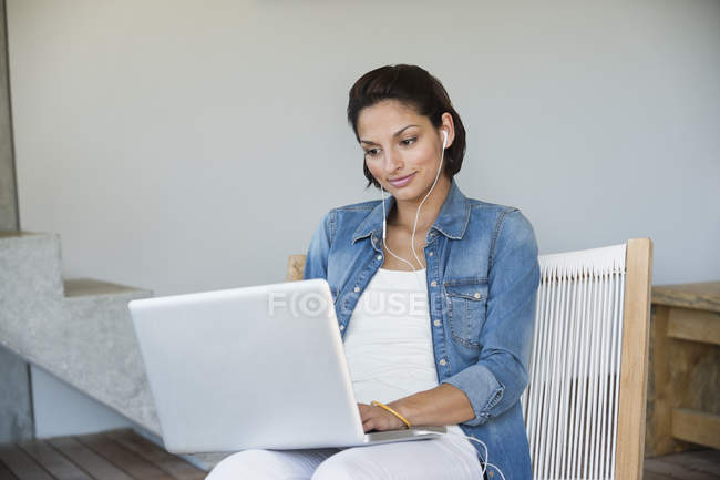 Жінка використовує ноутбук і слухає музику, сидячи на стільці — стокове фото