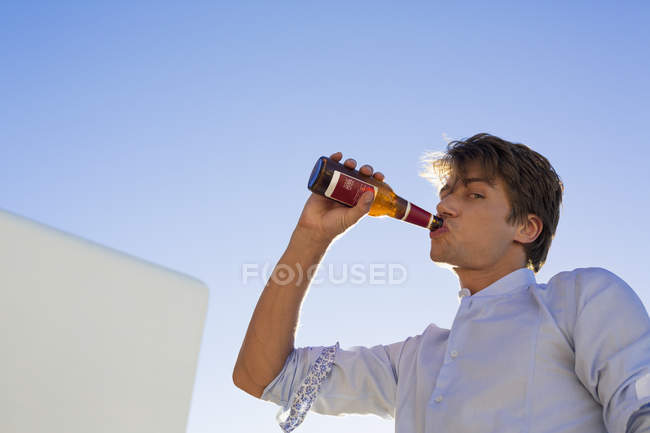 Молодой человек пьет пиво на фоне голубого неба — стоковое фото