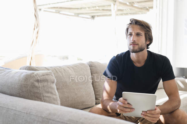 Jovem homem pensativo segurando tablet digital enquanto sentado no sofá e olhando para longe — Fotografia de Stock