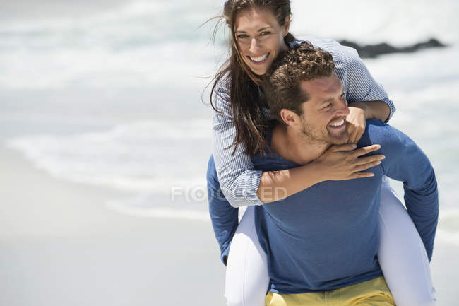 Cheerful man giving piggyback ride to wife on beach — Stock Photo