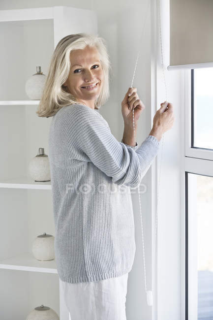 Старша жінка регулює віконну завісу вдома і дивиться на камеру — стокове фото