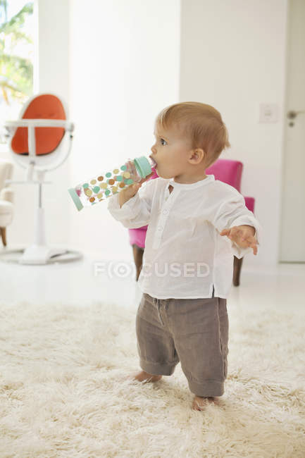 Дитячий хлопчик п'є воду з пляшки, стоячи на білому пухнастому килимі вдома — стокове фото