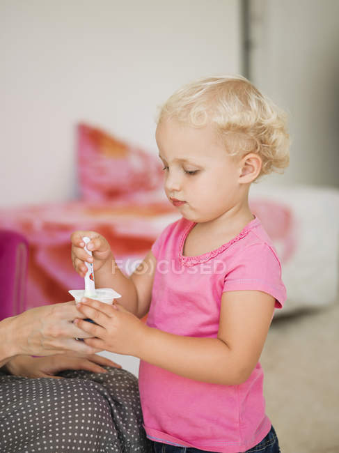 Cute blond girl eating yogurt from mother hand at home — Stock Photo