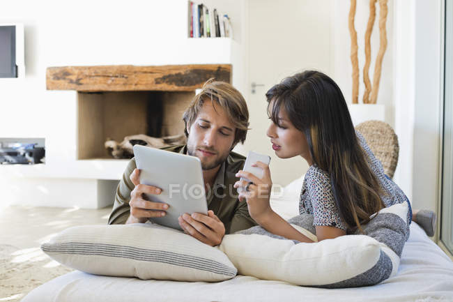 Casal deitado na cama e usando aparelhos eletrônicos — Fotografia de Stock