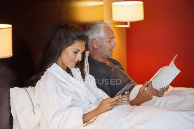 Mulher usando tablet digital com marido ler livro no quarto de hotel — Fotografia de Stock