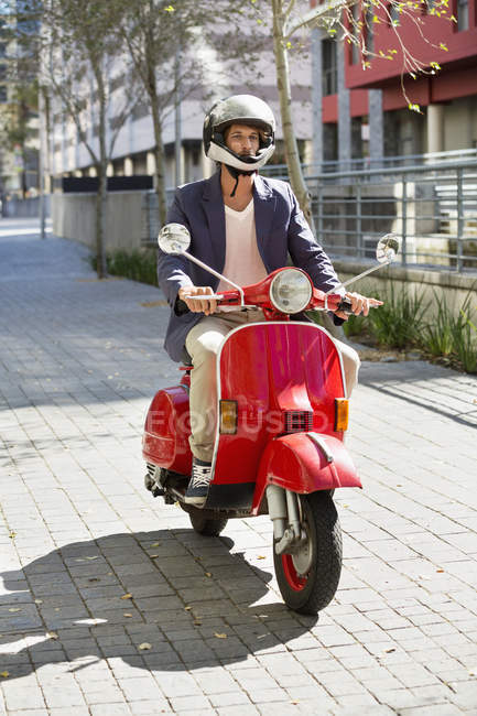 Человек в шлеме едет на красном скутере вниз по улице — стоковое фото