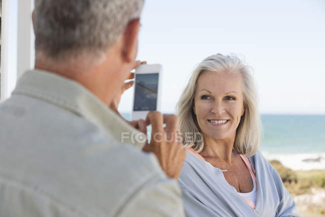 Man taking photo of wife with cell phone on beach — Stock Photo