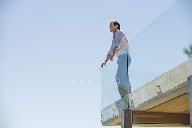 Pensativo hombre de pie en la terraza y mirando hacia otro lado contra el cielo azul - foto de stock