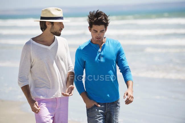 Расслабленные мужчины, идущие по пляжу с волнистым морем — стоковое фото