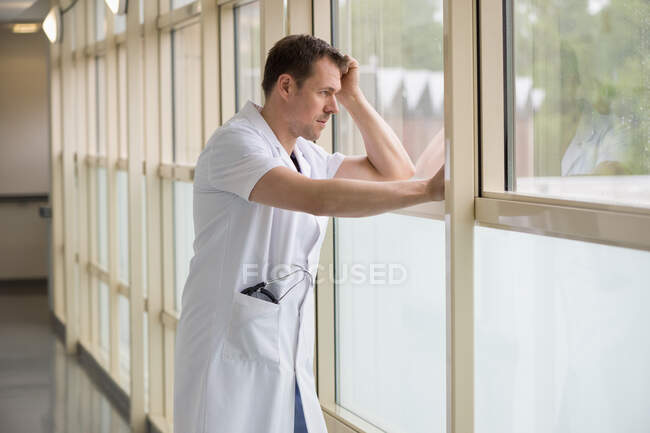 Médico varón de pie en el hospital mirando por la ventana - foto de stock