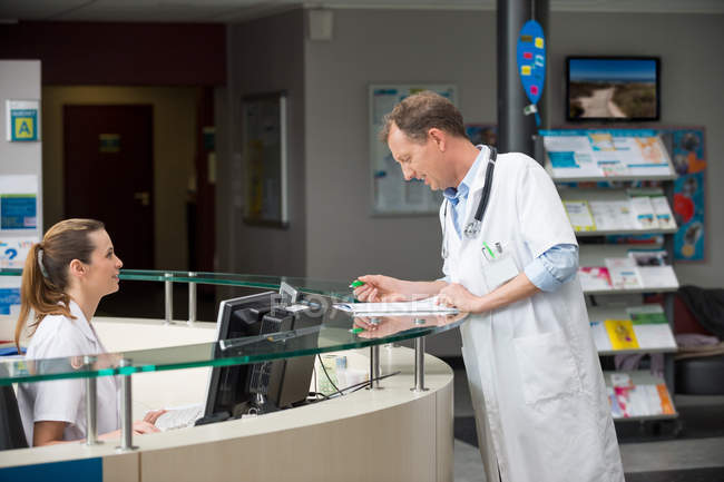 Médico masculino discutiendo papel con recepcionista en recepción del hospital - foto de stock