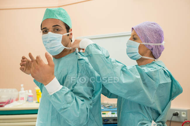 Cirujano femenino ayudando a un cirujano masculino a ponerse ropa protectora - foto de stock