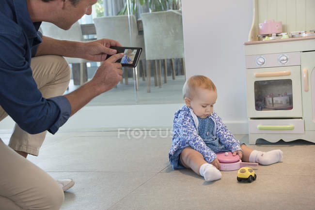Зрелый мужчина фотографирует маленькую дочь, играющую с игрушками на полу дома — стоковое фото