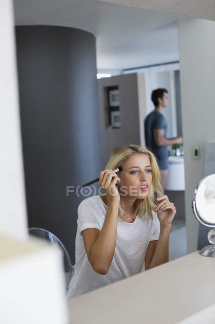 Молодая женщина наносит макияж на лицо с мужем на заднем плане в ванной комнате — стоковое фото