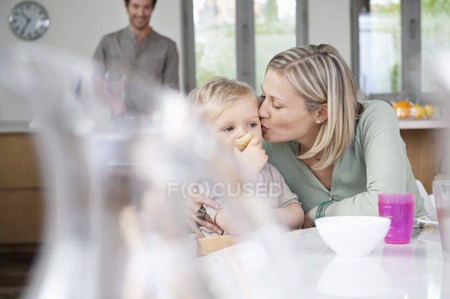 Woman eating breakfast with little son in kitchen with husband on background — Stock Photo