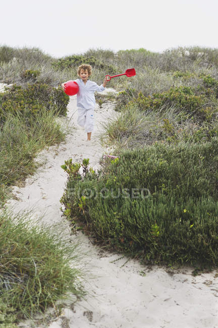 Menino alegre correndo no caminho arenoso na costa com pá vermelha e bola — Fotografia de Stock