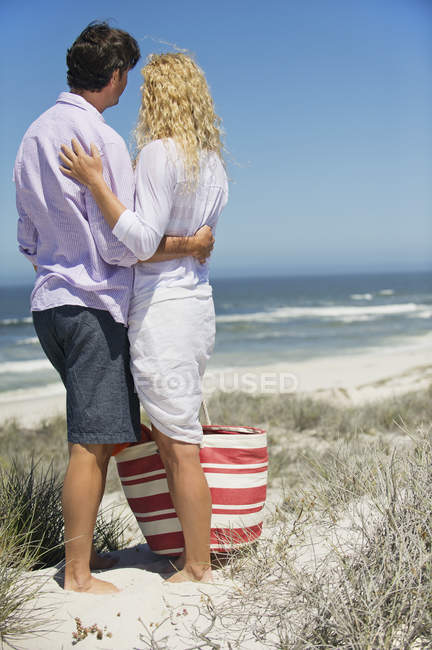 Vista trasera de pareja romántica abrazando y mirando a la vista en la playa - foto de stock