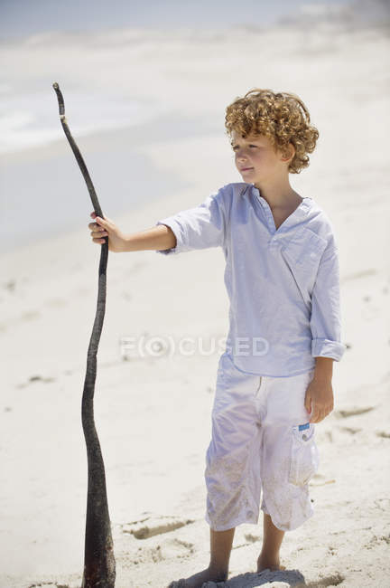 Menino segurando pau de madeira na praia de areia — Fotografia de Stock