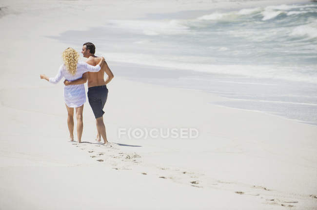 Vista trasera de abrazar pareja caminando en la playa - foto de stock