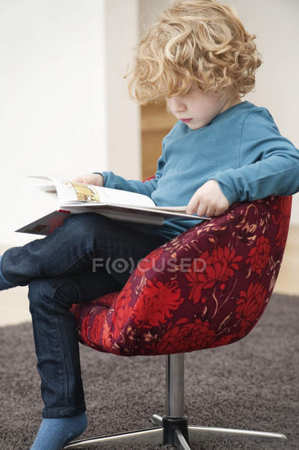 Carino ragazzo con i capelli biondi leggere un libro in poltrona a casa — Foto stock