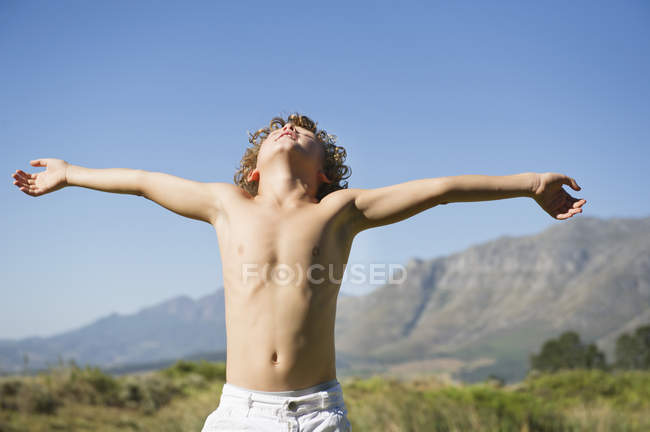 Shirtless little boy standing with eyes closed and arms outstretched against mountains — Stock Photo