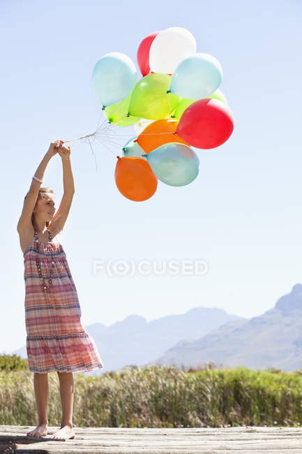 Girl in checkered dress playing with colorful balloons on pier in nature — Stock Photo