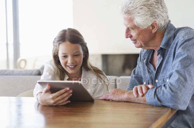 Chica usando tableta digital con el abuelo sentado en el escritorio - foto de stock
