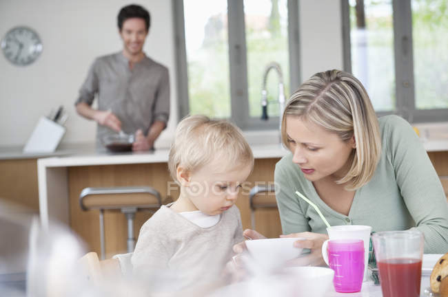 Mãe e pequeno filho sentado à mesa de café da manhã na cozinha com o marido no fundo — Fotografia de Stock