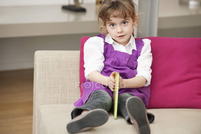 Portrait of cute little girl holding a book while sitting on sofa — Stock Photo
