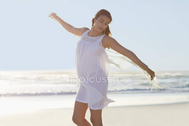 Young woman in white summer dress standing on beach with arms outstretched — Stock Photo