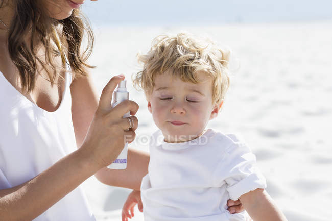 Женщина брызгает солнцезащитный крем на детское лицо на пляже — стоковое фото